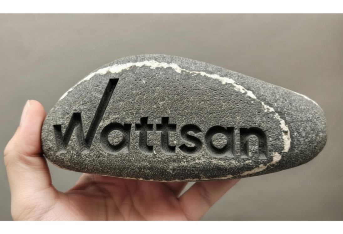 Рука держит серый камень с выгравированным словом "Wattson" жирными буквами